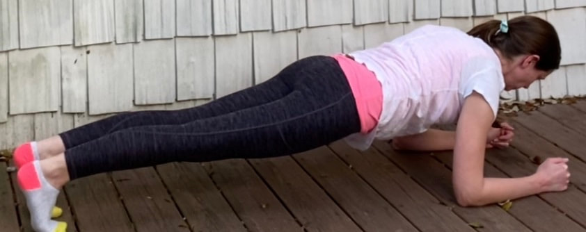 Picture of a female MotivateU fitness instructor completing a forearm plank pose outside on a brown wood deck with beige shingles in the background.