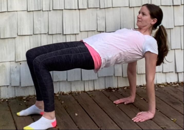 Picture of a female MotivateU fitness instructor completing a reverse table pose outside on a brown wood deck with beige shingles behind her, looking forward smiling.