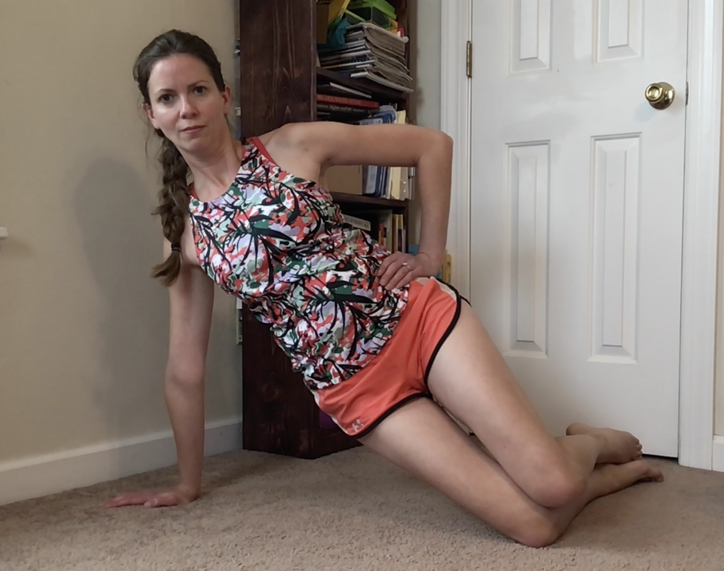 Picture of a female MotivateU fitness instructor completing a modified side plank pose on a carpet with a white closet door and brown bookshelf behind her, she is looking at the camera smiling.